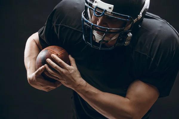 Jugador de fútbol americano con casco y armadura con una pelota contra la pared negra —  Fotos de Stock