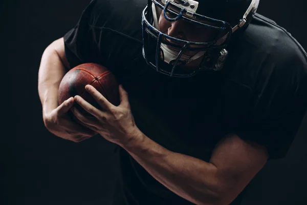 Jugador de fútbol americano con casco y armadura con una pelota contra la pared negra — Foto de Stock