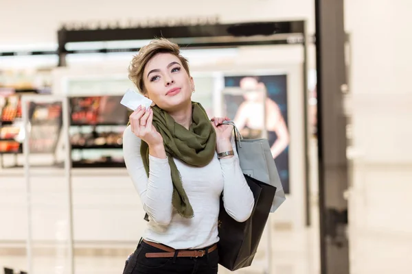 Mujer rubia con bolsas de compras en una mano y tarjeta de crédito en otra en el centro comercial — Foto de Stock