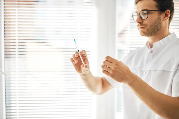 Mann in Arztuniform betrachtet Spritze vor Injektion am Arbeitsplatz — Stockfoto