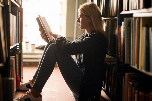 Young girl sitting on the floor in library between bookshelves, reading books.