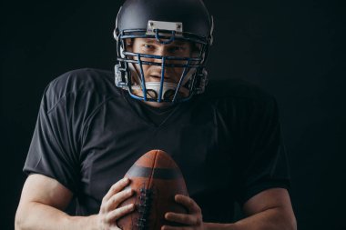 Siyah arka planda topu olan siyah spor giyimli Amerikalı bir futbolcu.