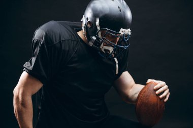 Siyah arka planda topu olan siyah spor giyimli Amerikalı bir futbolcu.