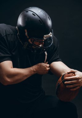 Siyah arka planda topu olan siyah spor giyimli Amerikalı bir futbolcu.