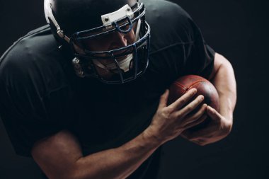 Kasklı ve zırhlı Amerikan futbolcu siyah duvara karşı top taşıyor.