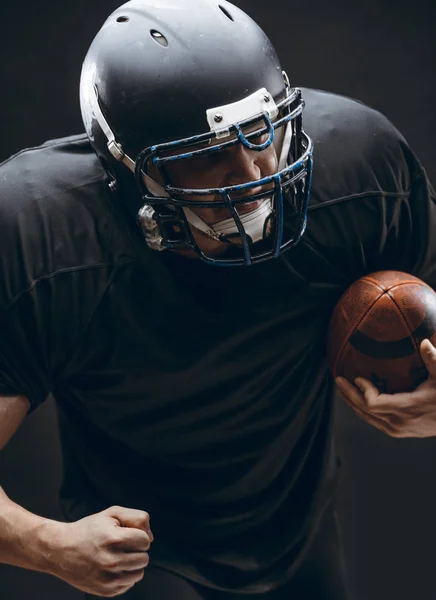 Jugador de fútbol americano con casco y armadura con una pelota contra la pared negra —  Fotos de Stock