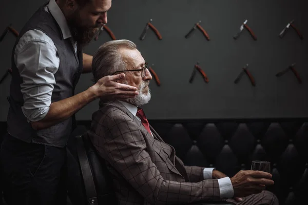 Bearded old businessman sitting at barber shop in chair choosing haircut design — Stock Photo, Image