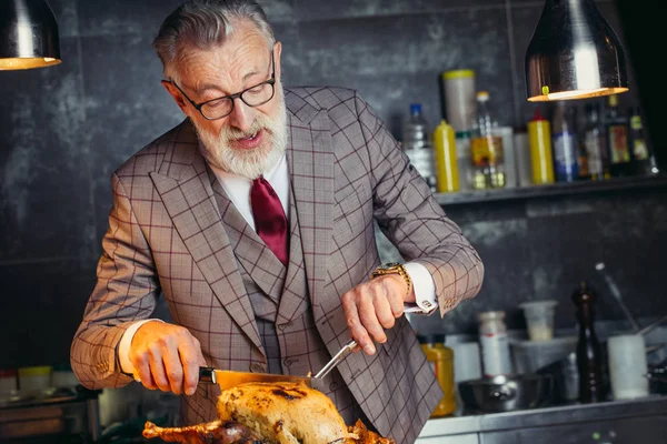 Hombre guapo de edad avanzada en traje formal costoso recortando pollo asado — Foto de Stock
