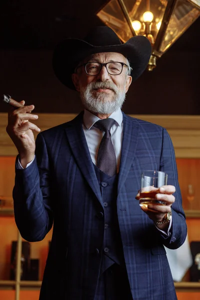 Stijlvolle oude man met brede hoed en rijke blauwe mannen pak rook — Stockfoto