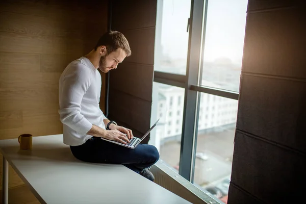 Bebaarde jonge zakenman in witte pullover werkt op computer. — Stockfoto