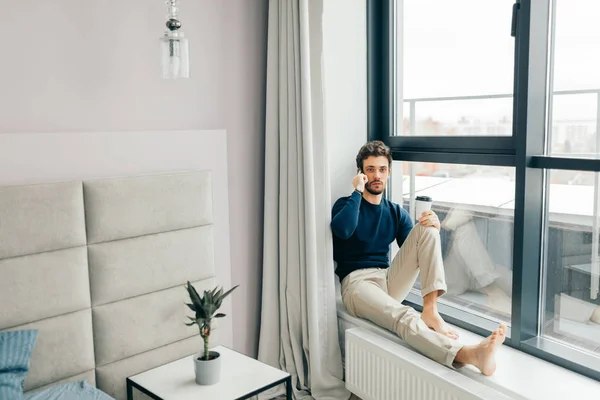 Schöner Mann sitzt mit Smartphone in der Hand auf der Fensterbank. — Stockfoto