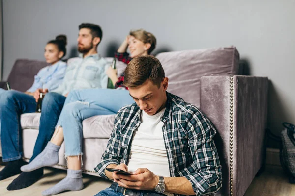 young man in checked shirt is sending sms while his friends are watching TV