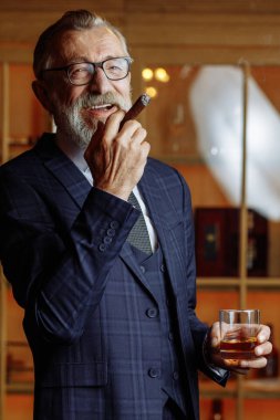 Resmi takım elbiseli yaşlı bir işadamı viski ve puro içiyor.