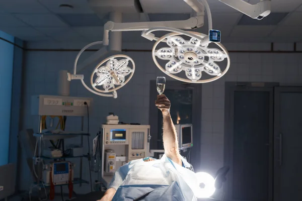Ironiczne zdjęcie pacjenta na kanapie operacyjnej podnosi rękę szampanem — Zdjęcie stockowe