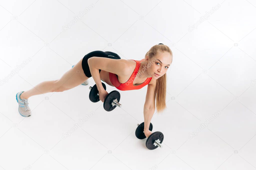 positive blonde sportswoman is training with dumbbells and looking at the camera. full length photo.