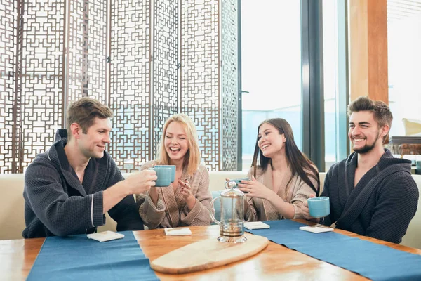 Relaxed diverse friends in bathrobes sitting at lounge zone of bathhouse