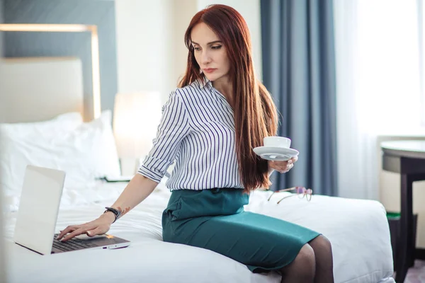 Femme aux cheveux roux en tenue de cérémonie prenant un café et travaillant sur un ordinateur portable au lit . — Photo