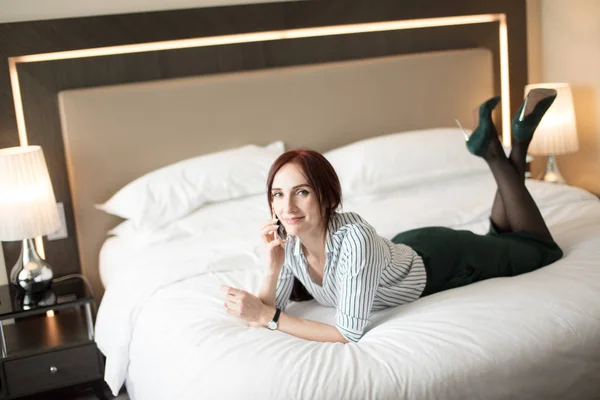 Elegant stylish businesswoman wearing high-heeled shoes lying on bed in hotel
