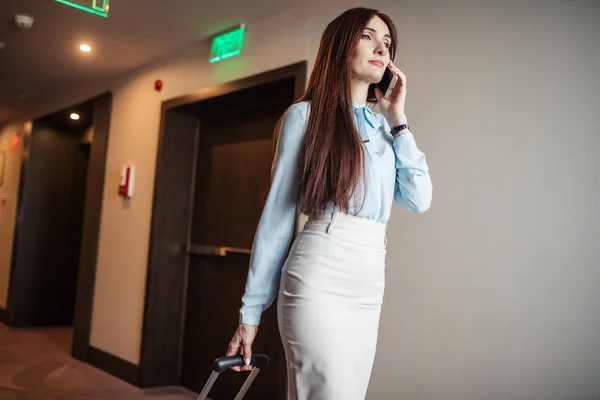 Mujer elegante hablando por teléfono inteligente mientras sale del hotel con la maleta . — Foto de Stock
