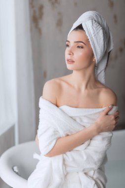 Bornozlu kahverengi saçlı kadın, banyo tüpünün yanında çıplak omuzlarıyla poz veriyor.