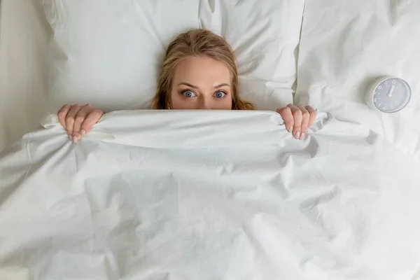 Pleasant blonde girl hiding under the blanket and looking at the camera — Stock Photo, Image