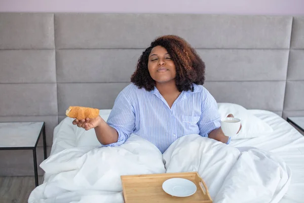 Mulher afro-americana gosta de café da manhã enquanto se senta na cama — Fotografia de Stock
