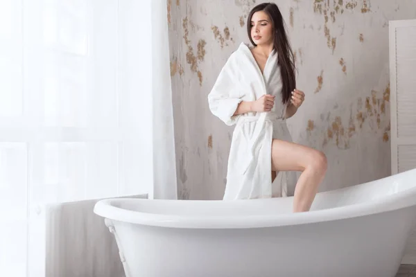 Fiatal nő fehér fürdőköpenyben ül a szabadon álló fehér fürdő mellett. — Stock Fotó