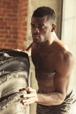 Athletic african american man exercising with tyre and looking away in gym clipart