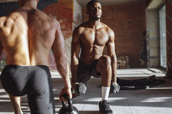 Two multiracial male athletes making lunges with weight in indoor workout