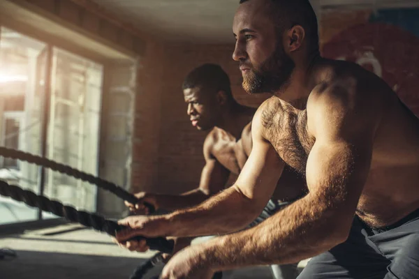 Dos deportistas de pecho desnudos multiétnicos haciendo ejercicio con cuerdas de batalla en el gimnasio . — Foto de Stock