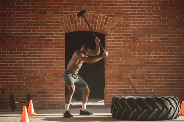 Muscular african man hitting wheel tire with sledgehammer against brick wall