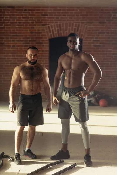 Dos deportistas de pecho desnudos multiétnicos haciendo ejercicio con cuerdas de batalla en el gimnasio . —  Fotos de Stock