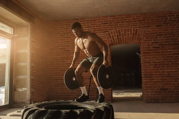 Shirtless african man with barbell plates jumps in air on truck tire in gym