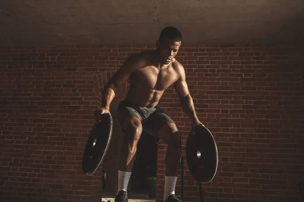 Shirtless african man with barbell plates jumps in air on truck tire in gym