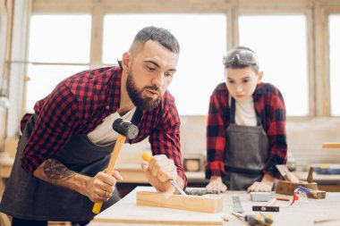 Sakallı ve gözlüklü bir çocuk marangozhanede çalışıyor..