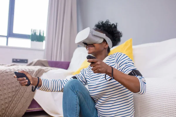 Mulher africana assistindo vídeo usando óculos VR com controle remoto em casa . — Fotografia de Stock
