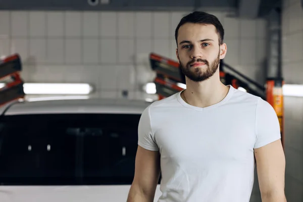 attractive bearded guy in a white T-shirt standing in front of the car