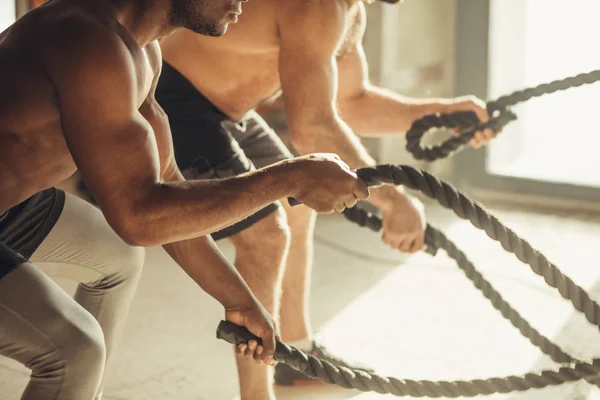 Dos deportistas de pecho desnudos multiétnicos haciendo ejercicio con cuerdas de batalla en el gimnasio . — Foto de Stock