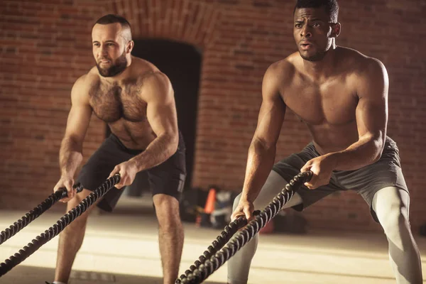 Twee multi-etnisch blote borst sporters trainen met de slag bij touwen op de sportschool. — Stockfoto