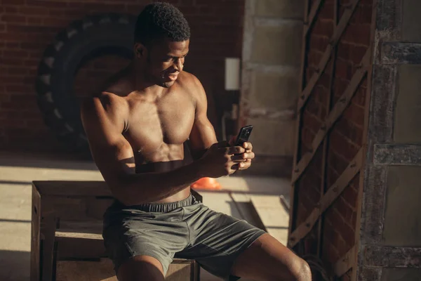 side view of shirtless african american athlete resting and using smartphone on tire at gym