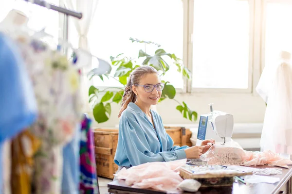 Jonge vrouwelijke kleermaker zit op werkplek en kiest de kleur van de draden in atelier — Stockfoto