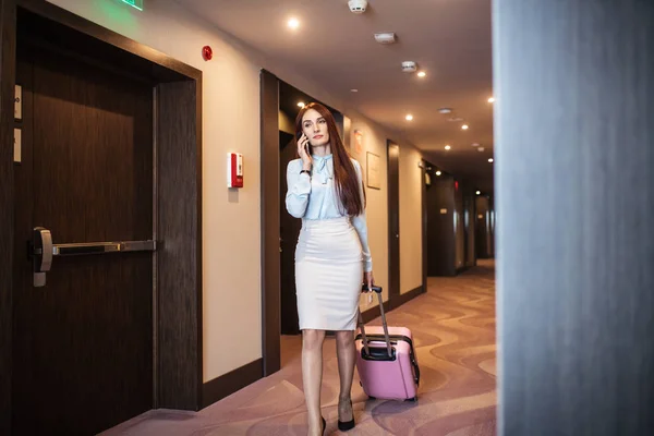 Elegant woman speaking on smart phone while leaving hotel with suitcase. — Stock Photo, Image