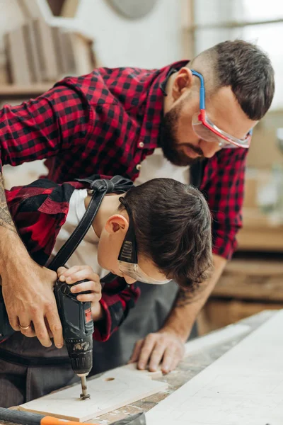 Otec a malý syn pracují s vrtačkou perforovanou dřevěnou deskou v dílně — Stock fotografie