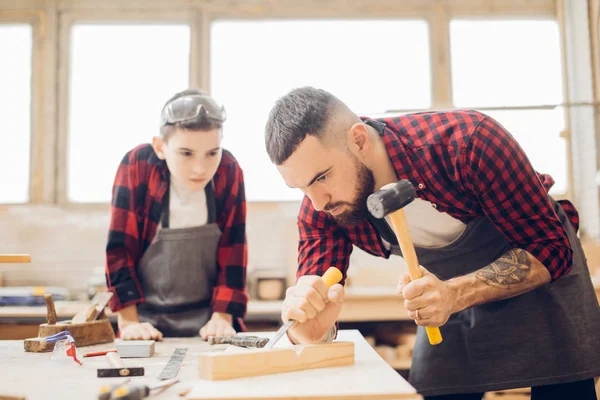 Bearded man with a boy in safety glasses works in a carpentry shop.