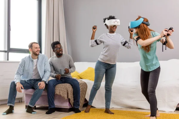 Dos jóvenes multirraciales bailando con gafas de realidad virtual mientras sus novios se sientan en el sofá y sonríen — Foto de Stock