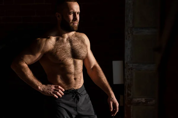 Сильный симпатичный спортсмен без рубашки позирует возле темной кирпичной стены — стоковое фото