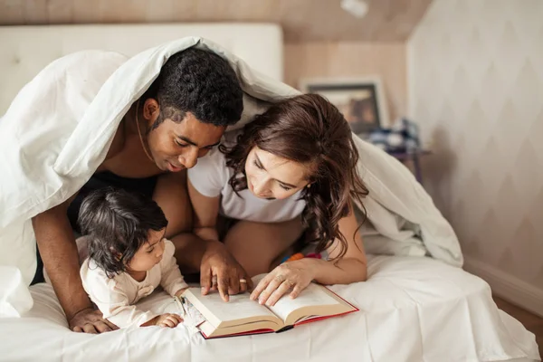 Šťastní rodiče s knihou veselé dítě, zatímco krycí s dekou — Stock fotografie