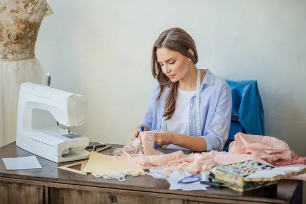 Schöne fokussierte junge Modedesignerin schneidet rosa Spitzenstoff in der Werkstatt — Stockfoto