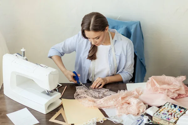 Schöne fokussierte junge Modedesignerin schneidet rosa Spitzenstoff in der Werkstatt — Stockfoto