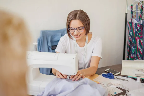 Joven modista sonriente cose ropa en una máquina de coser en su taller —  Fotos de Stock